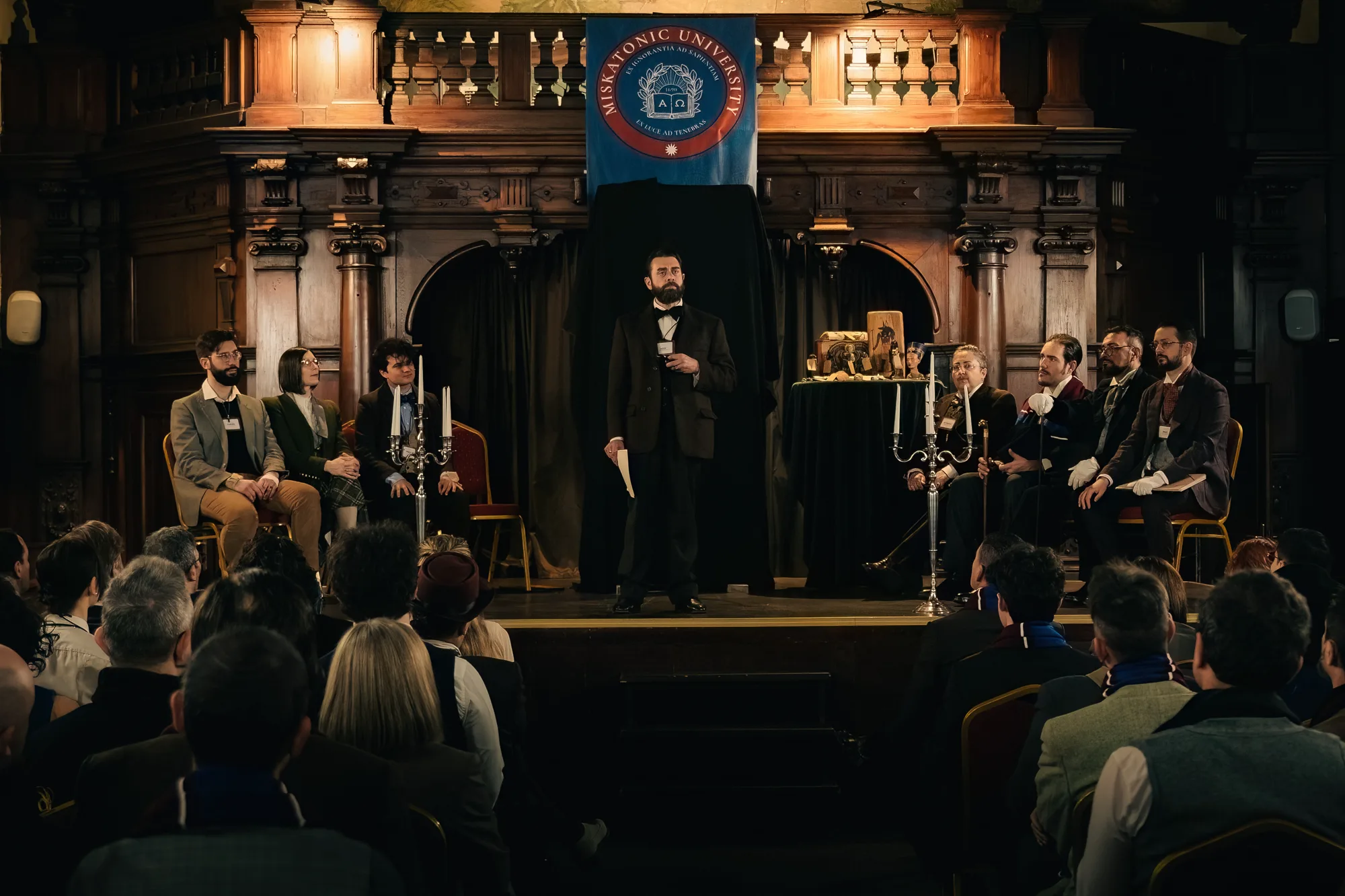 The Dean addresses the university from the stage, beneath its banner