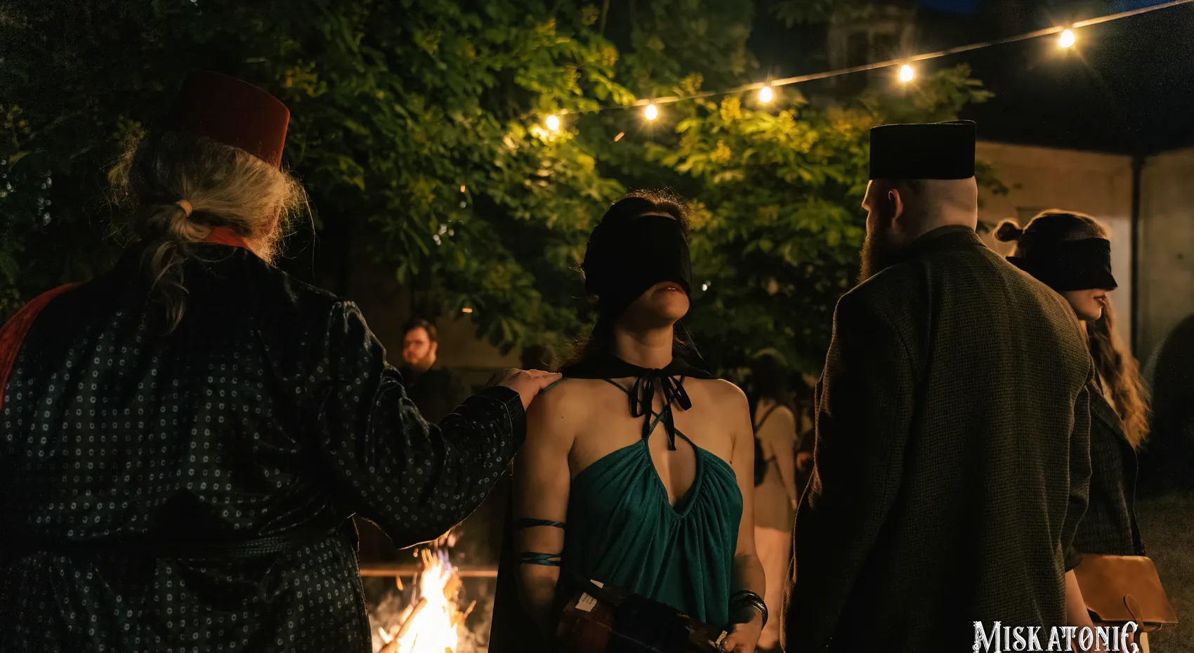 A blindfolded woman is led through the night, a fire behind